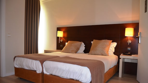 A double bed with two pillows, a bedside table with a lamp, and a large window with curtains.