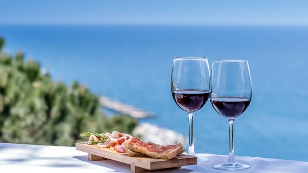 Une table sur laquelle est disposée une planche en bois recouverte de parts de pizza et deux verres de vin rouge, le tout sur fond de ciel bleu clair et d’océan.