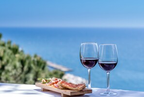 Una mesa con una tabla de pizza en porciones y dos copas de vino tinto, sobre un fondo de cielo azul despejado y mar.