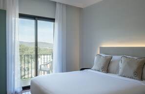 Habitación de hotel con una cama grande, un balcón con vistas a zonas verdes y colinas, y una decoración en tonos blancos.