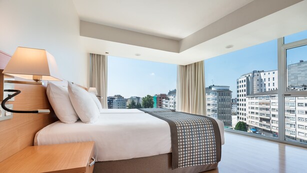 A modern hotel room with a large bed, a bedside table, a lamp, and a view of the cityscape through large windows.