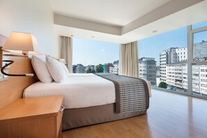 A modern hotel room with a large bed, a bedside table, a lamp, and a view of the cityscape through large windows. - Dorma Puerta de San Pedro (Lugo)