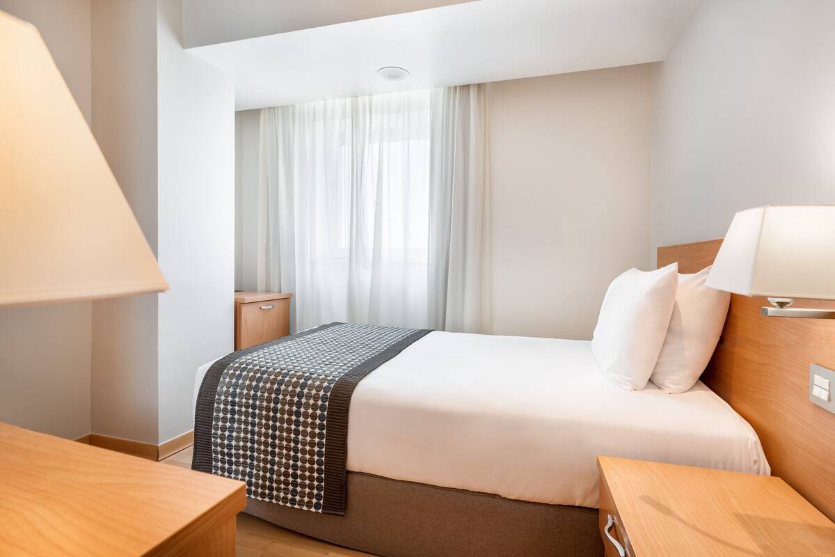 Habitación de hotel con cama, mesitas de noche, lámpara y ventana con cortinas.