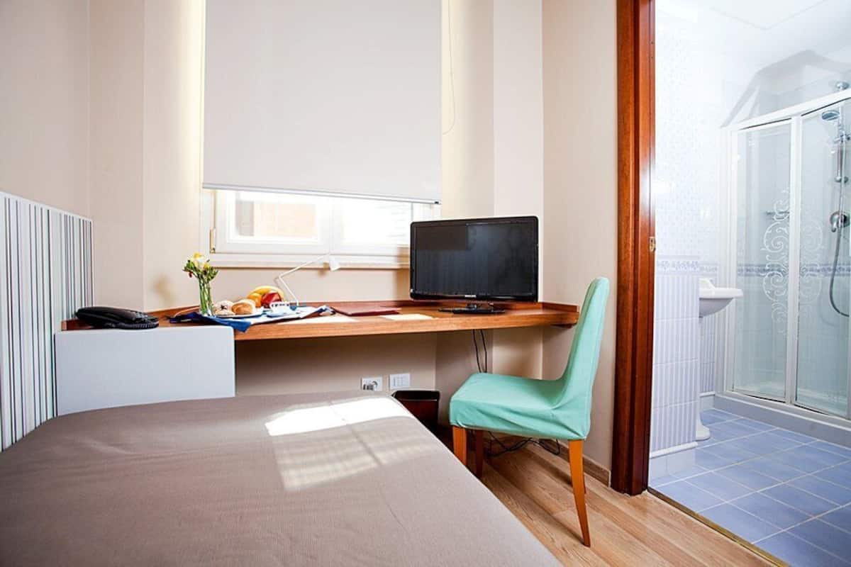 A hotel room with a bed, a desk with a computer, a chair, a window, and a shower area.