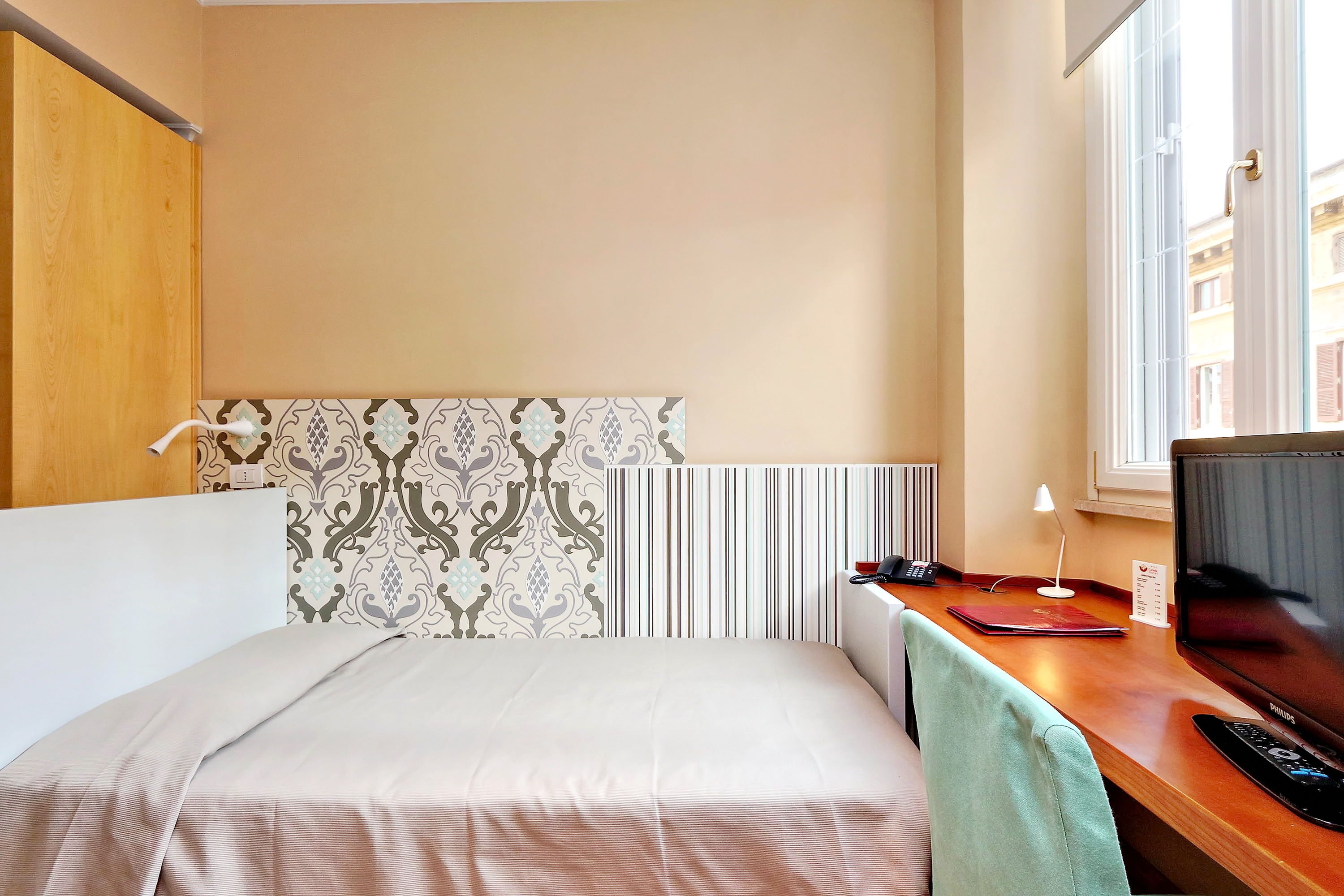 A hotel room with a bed, a desk with a computer, a lamp, and a window with a view of buildings.