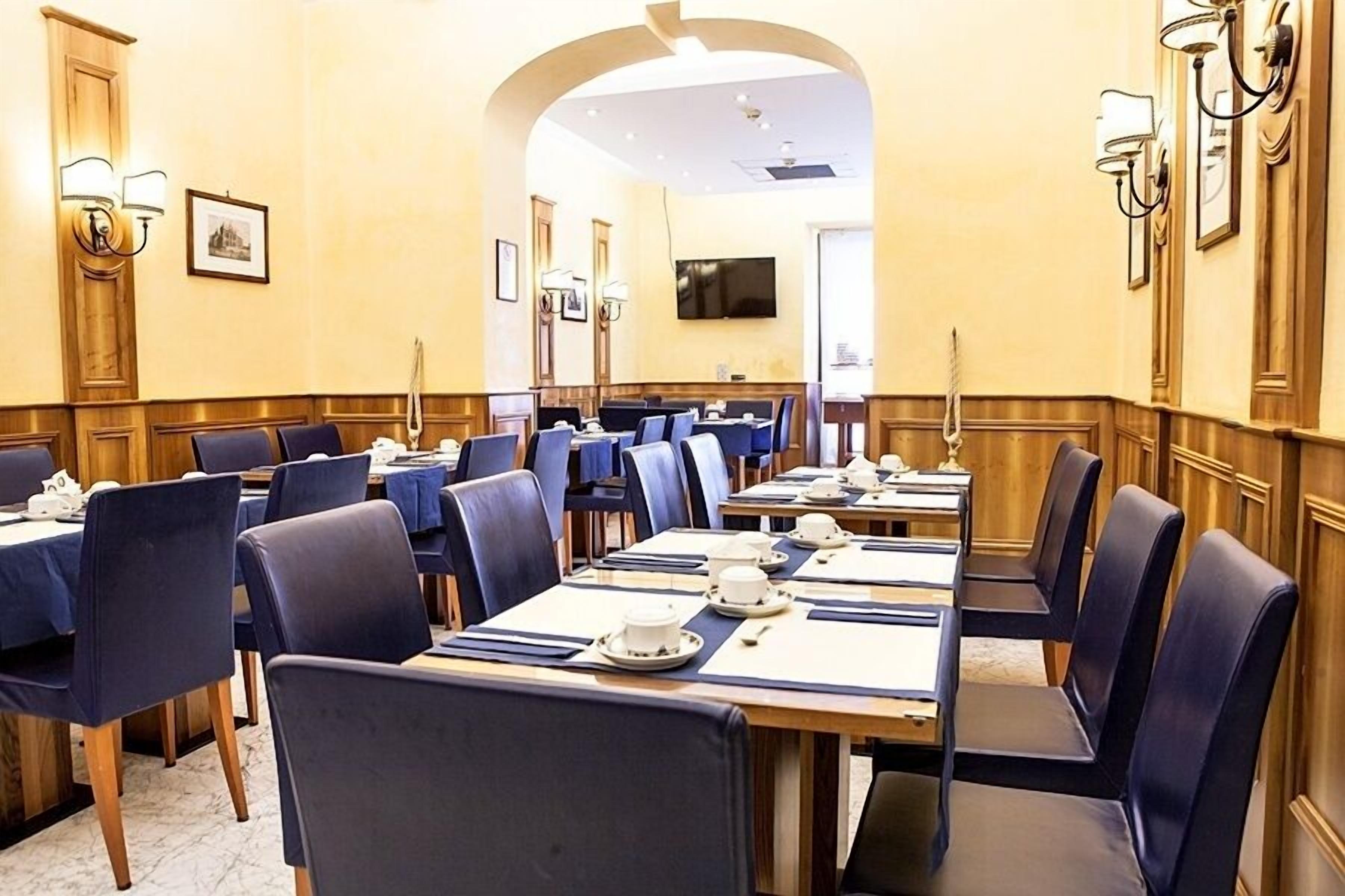 A hotel dining area with multiple tables and chairs, a television mounted on the wall, and framed pictures on the walls.