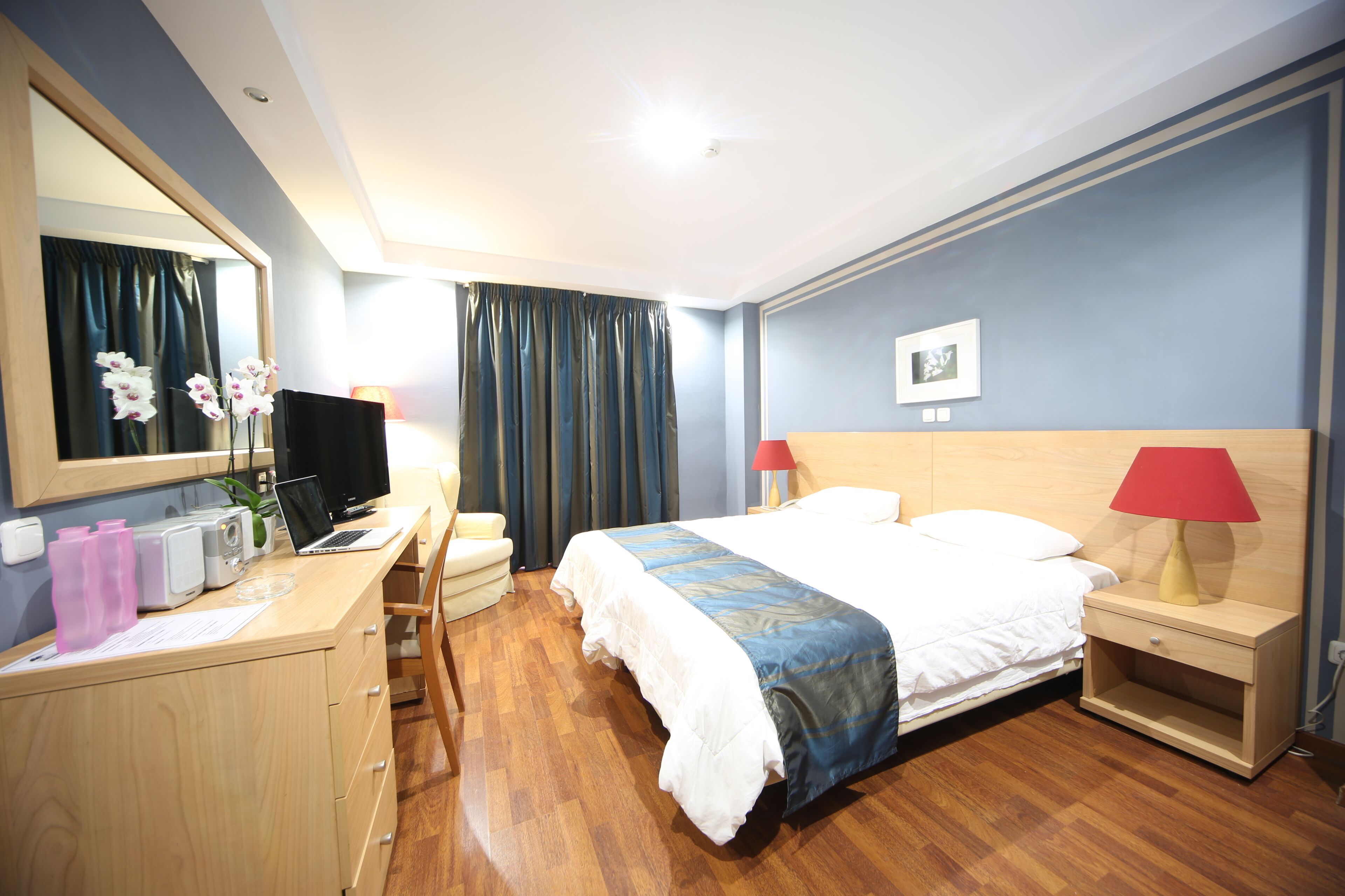 Hotelkamer met een bed, een bureau met een computer, een stoel, een lamp, een spiegel en een raam met gordijnen.