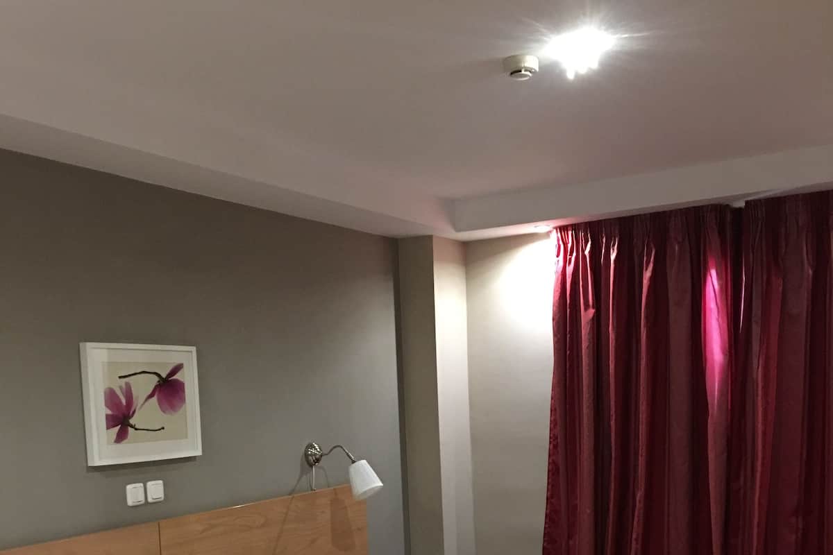 Een keurig opgemaakt bed met een beige dekbed en kussens, een bedlampje, een ingelijst foto op de muur en een rood gordijn.