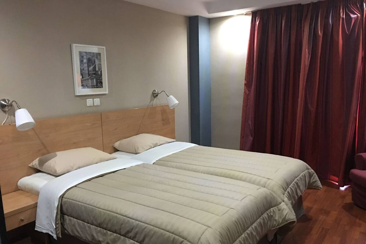 Een tweepersoonsbed met houten hoofdeinde, twee bedlampjes, een nachtkastje, een inlijsting foto aan de muur en een rood gordijn.