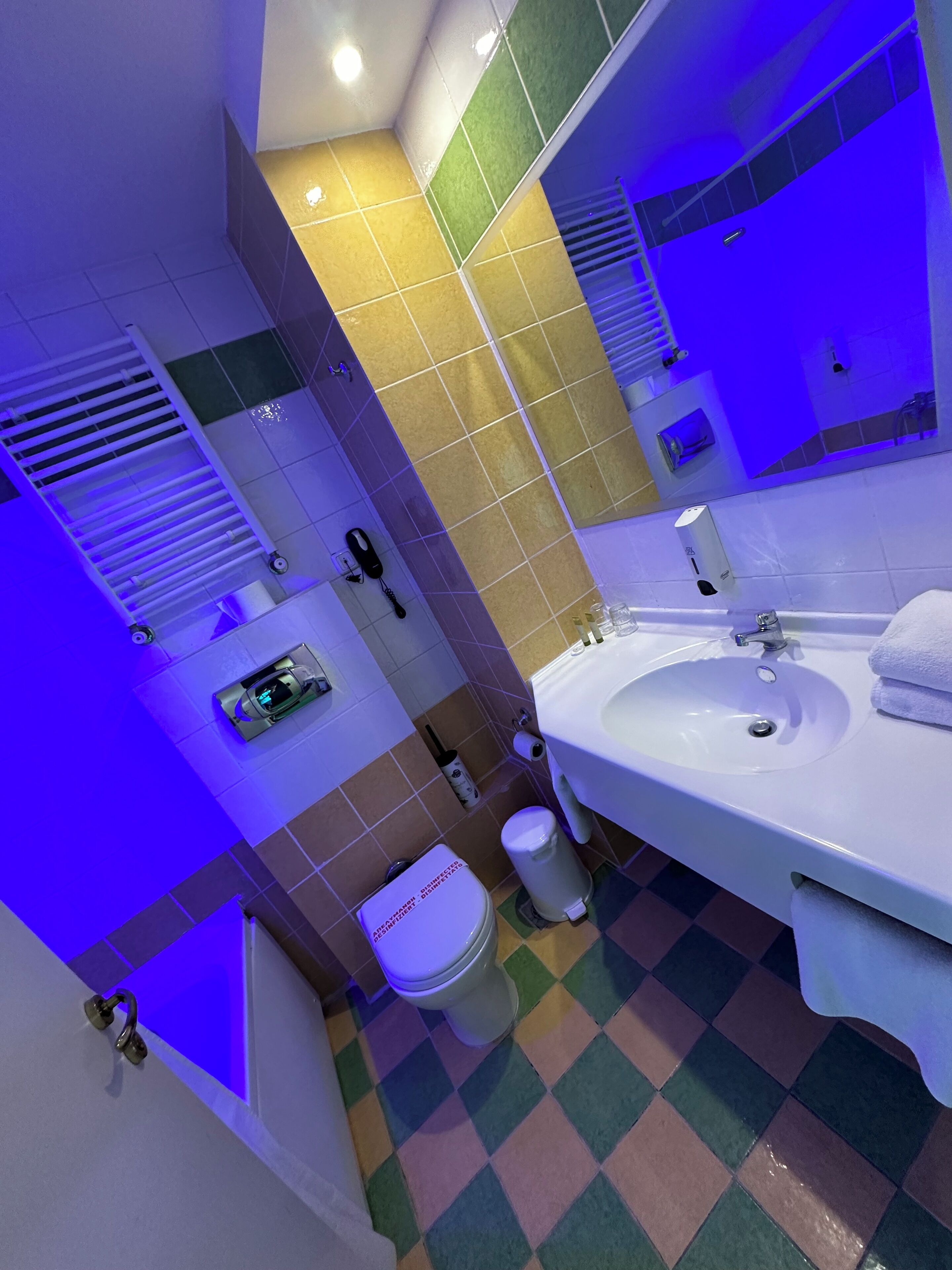 A modern bathroom with a large sink, toilet, and shower. It features a checkered floor pattern and a mirror with a blue light.