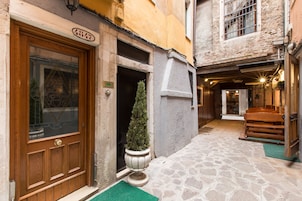 A narrow stone-paved alleyway leading to a wooden door with a green mat. A potted evergreen stands beside the door.