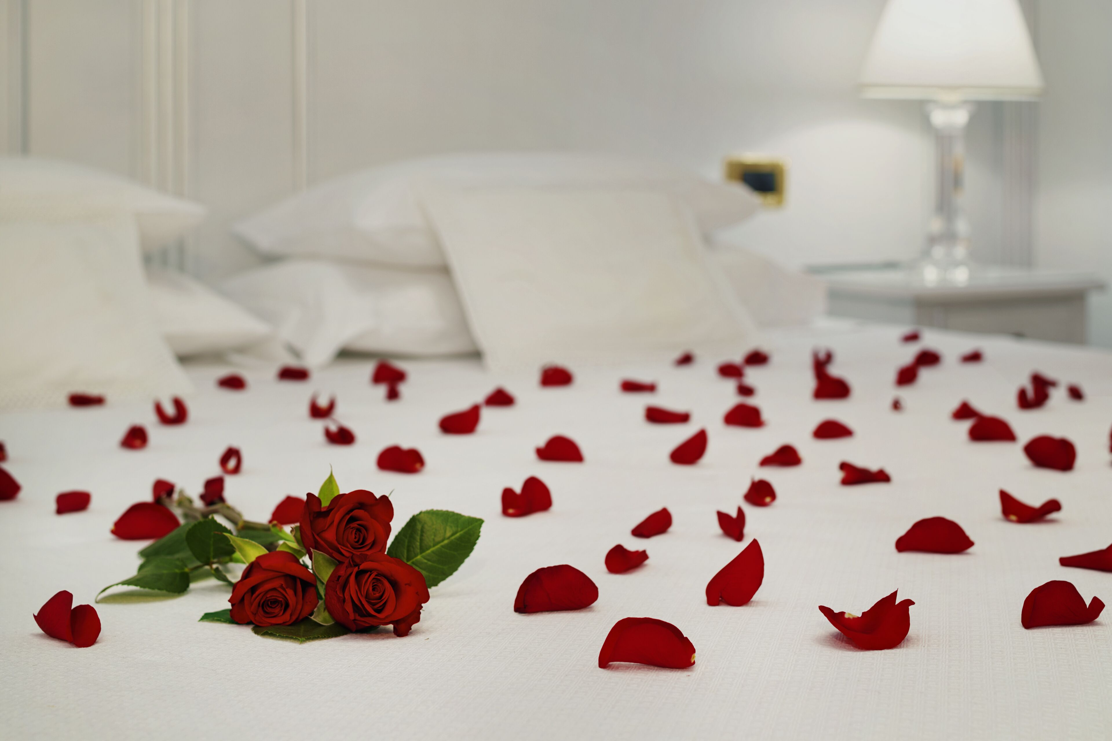 Una cama con pétalos y rosas, ropa de cama blanca y una mesita de noche con una lámpara.