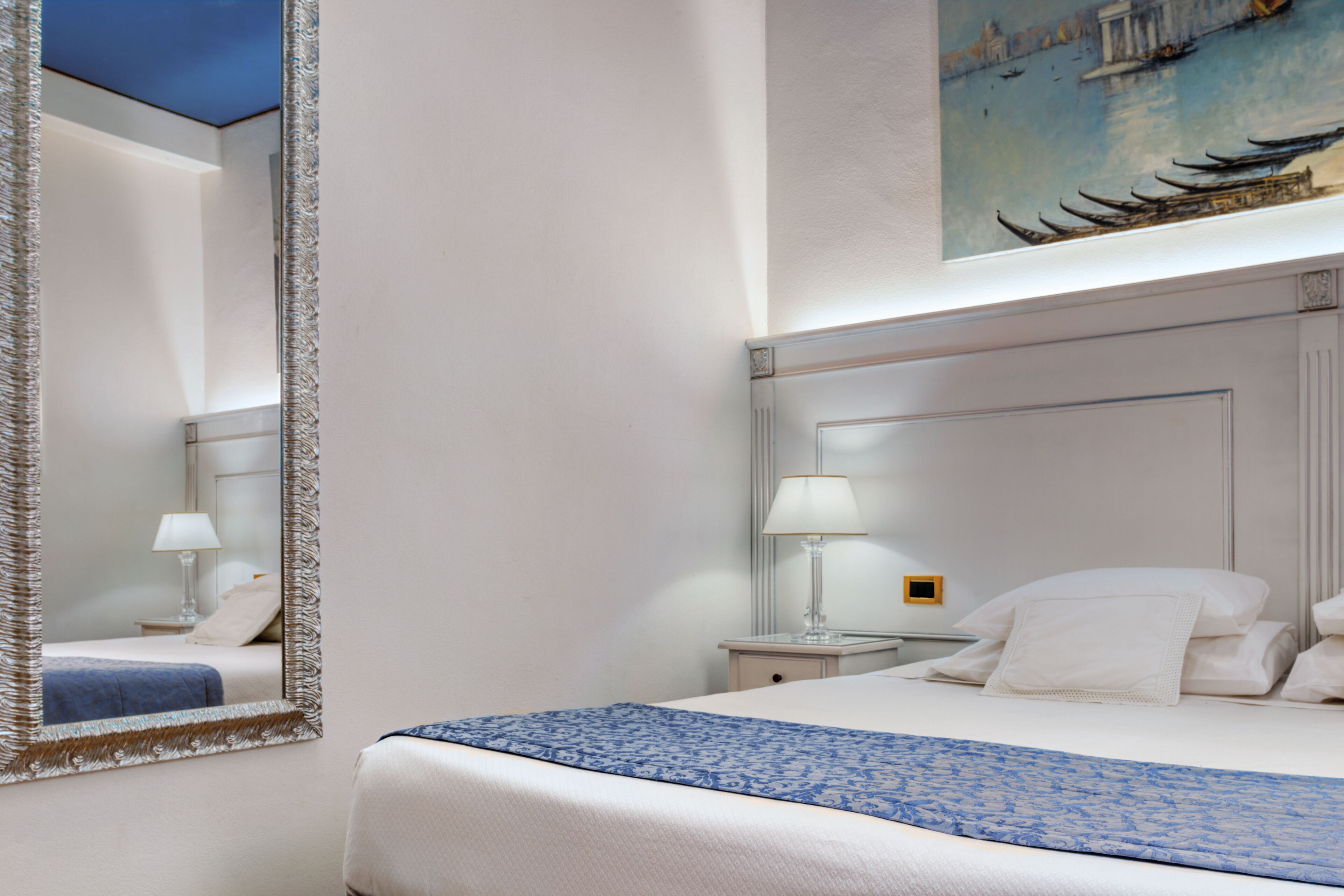 A hotel room with a bed, a nightstand with a lamp, a large mirror, and a painting of boats on the wall.