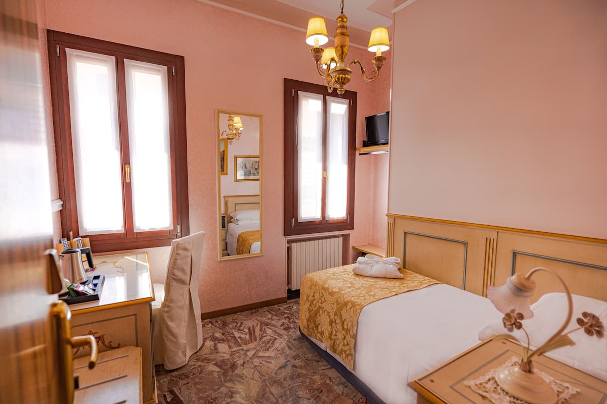 A hotel room with a bed, a desk with a telephone, a chair, a mirror, a television, and a chandelier.