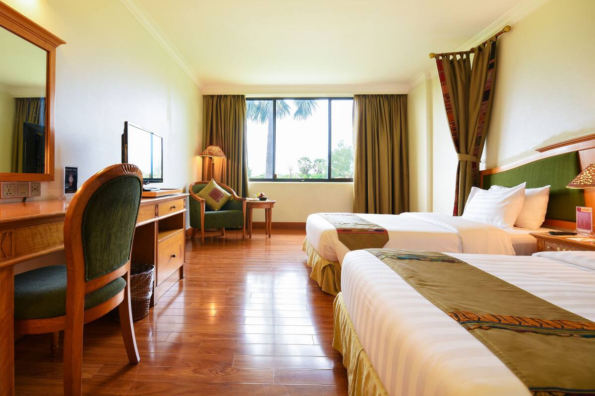 A hotel room with a wooden desk, two beds, a chair, a TV, and a window with curtains.
