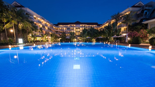 A large, well-lit swimming pool with a tiled deck, surrounded by palm trees and a multi-story hotel building with balconies.
