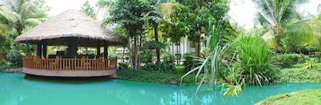 Un quiosco con techo de paja y baranda de madera, rodeado de exuberante vegetación y una piscina tranquila.