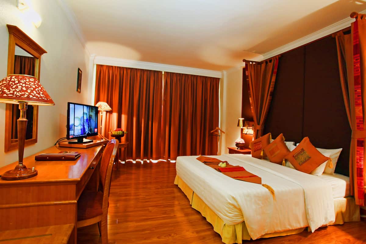 A hotel room with a wooden desk, a television, a bed with white bedding and orange pillows, and a large window with brown curtains.