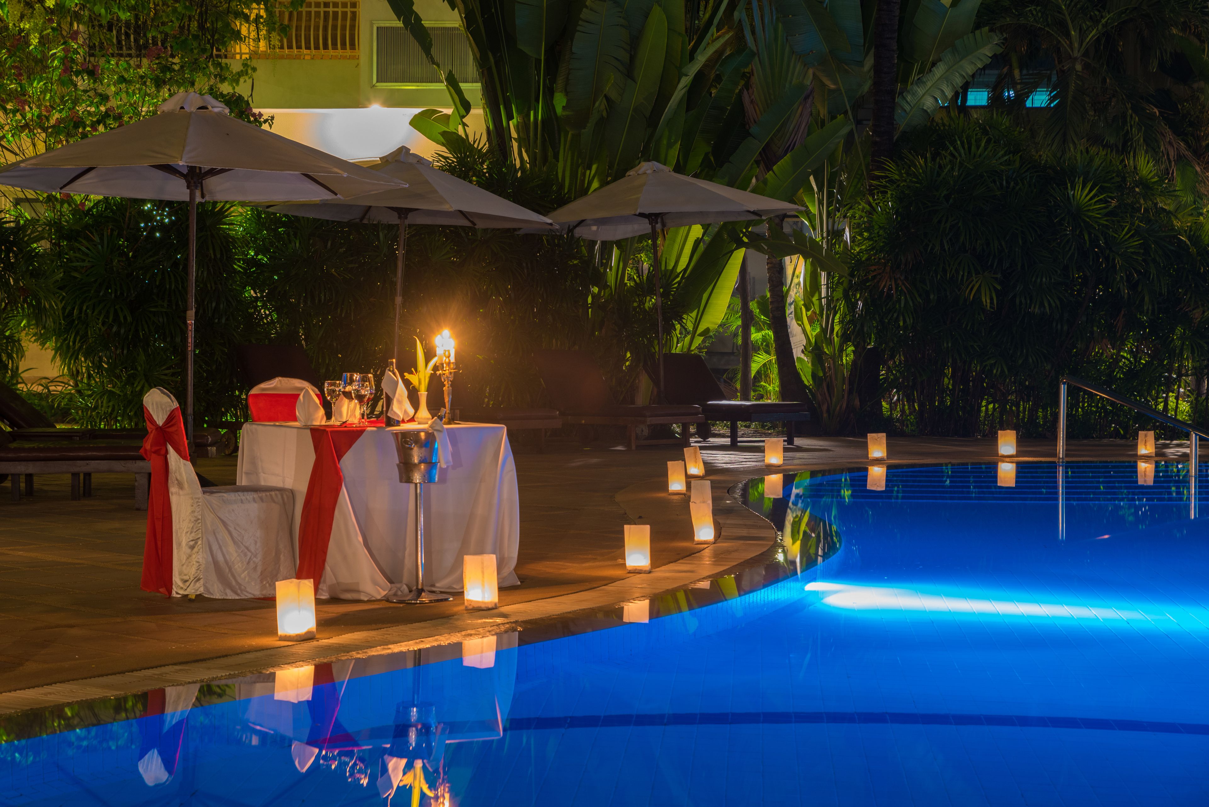 Zona junto a la piscina con mesa lista para comer, rodeada de sombrillas y velas.