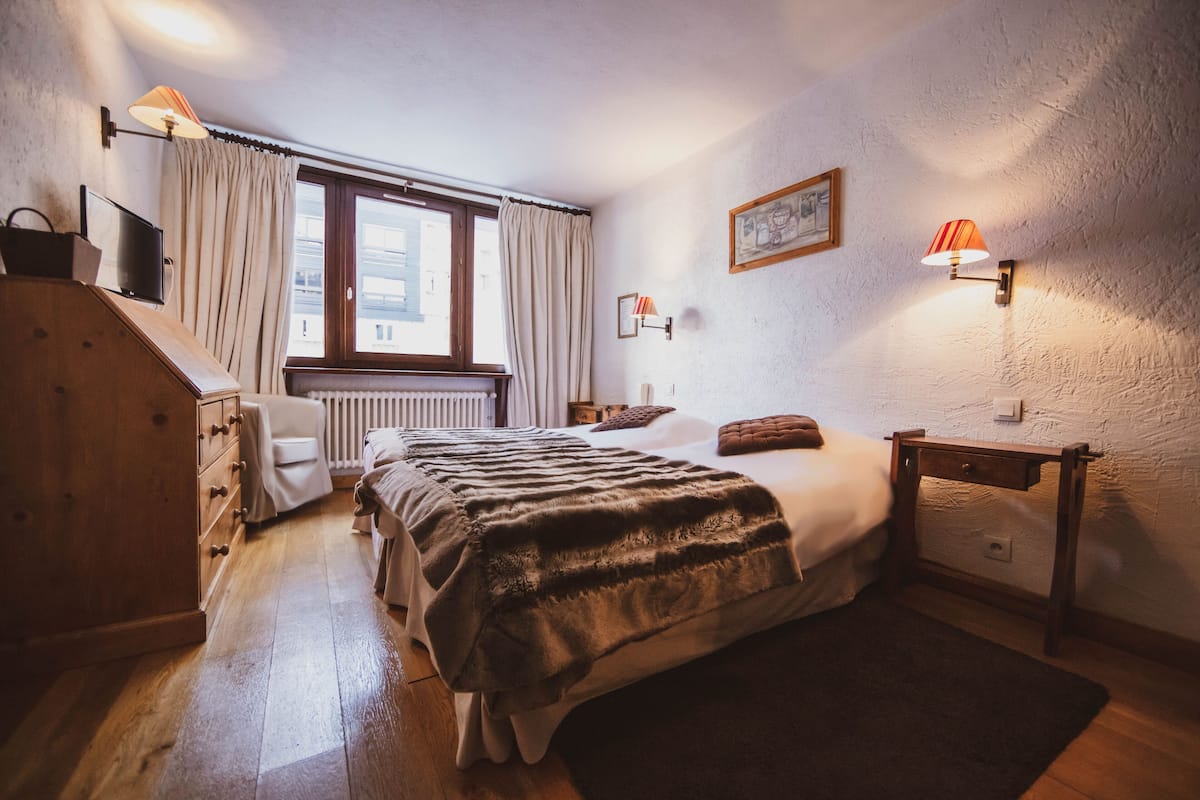 Un dormitorio con cama, cómoda de madera, silla, ventana con cortinas y lámparas fijadas a la pared.