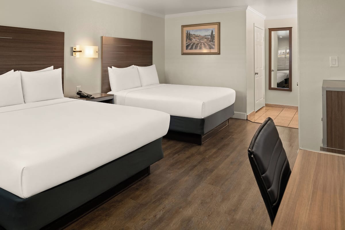 A hotel room with two beds, a desk, a chair, a nightstand, a painting, and a door leading to another room.