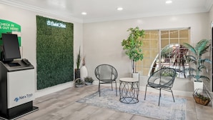 A reception area with a NexTap kiosk, a green wall with 'El Castillo Motel' signage, two chairs, a small table, and potted plants. - El Castell Motel (Monterey)