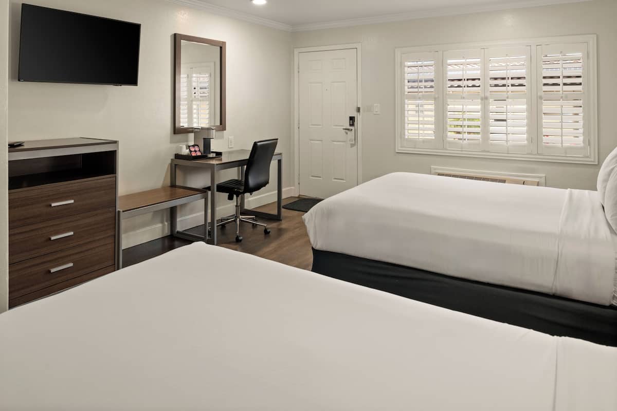 A hotel room with two beds, a desk with a chair, a TV, a mirror, a door, and a window with blinds.