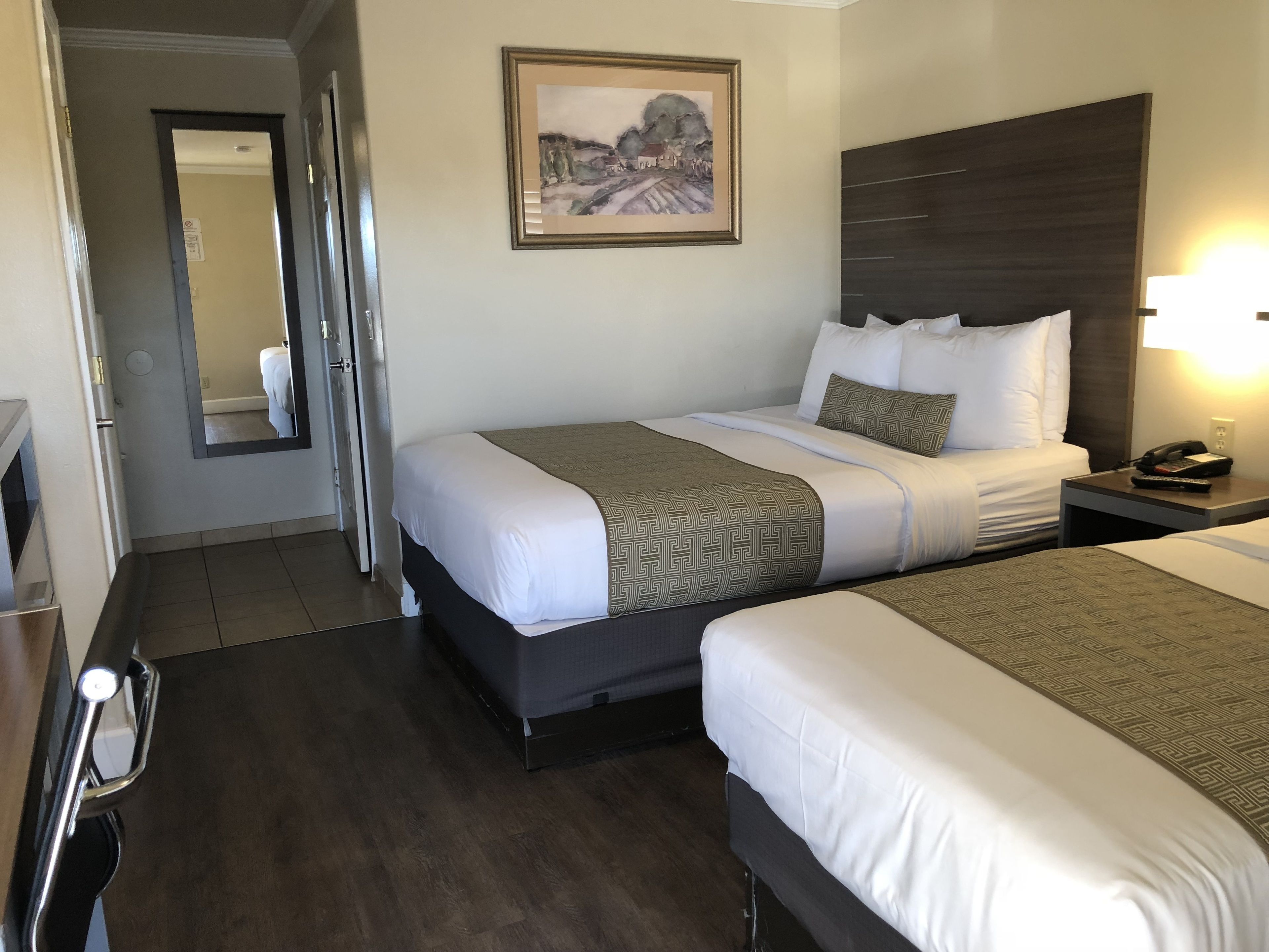 A hotel room with two beds, a large mirror, a painting on the wall, and a bedside table with a lamp.