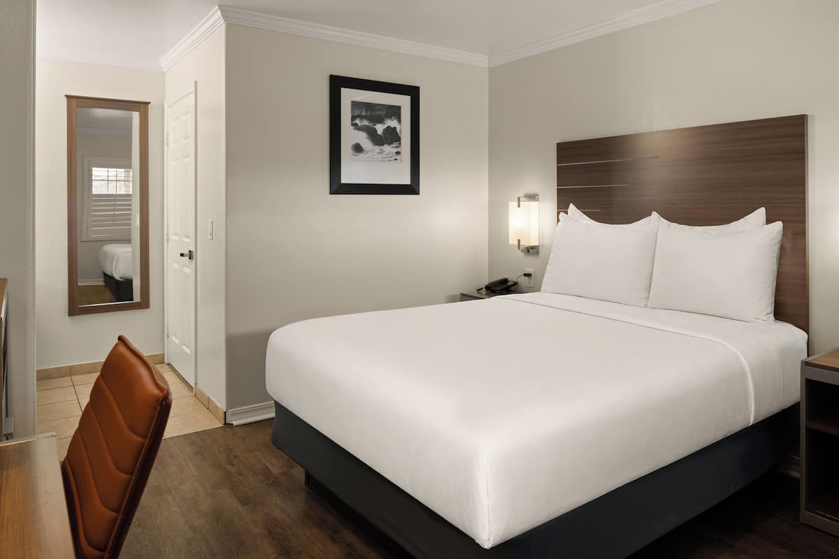 A hotel room with a large bed, a desk, a chair, a framed picture on the wall, and a nightstand with a lamp.