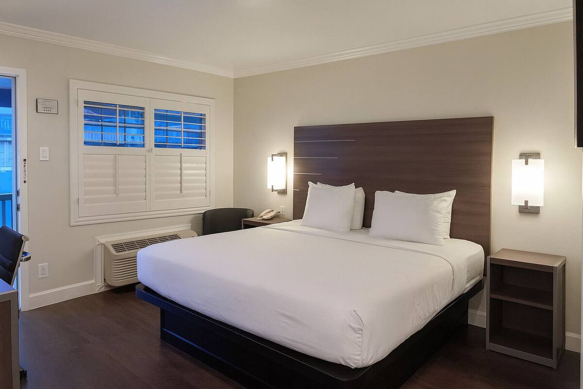 A hotel room with a large bed, a desk, a chair, a window with blinds, a wall-mounted light, and a small nightstand.
