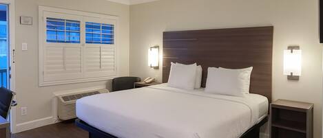 A hotel room with a large bed, a desk, a chair, a window with blinds, a wall-mounted light, and a small nightstand.