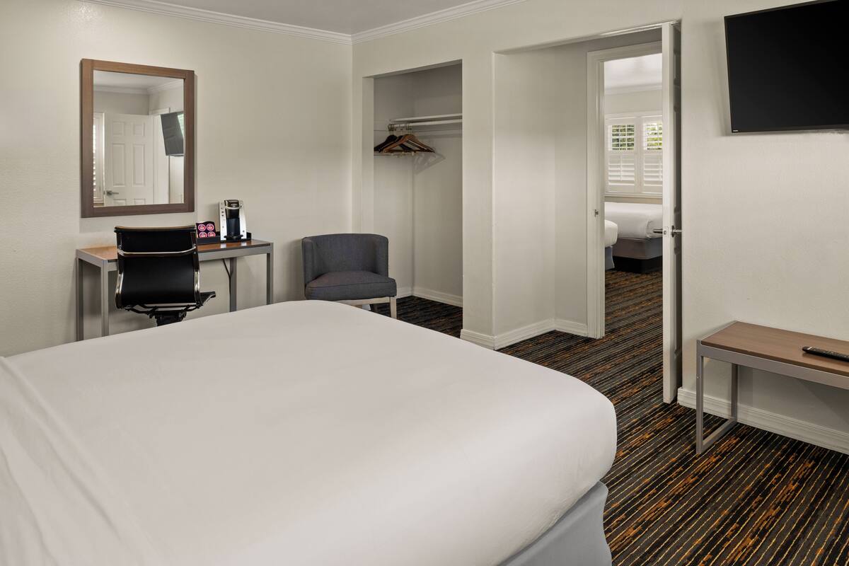 A hotel room with a bed, a desk with a chair, a coat rack, a television, and a door leading to another room.