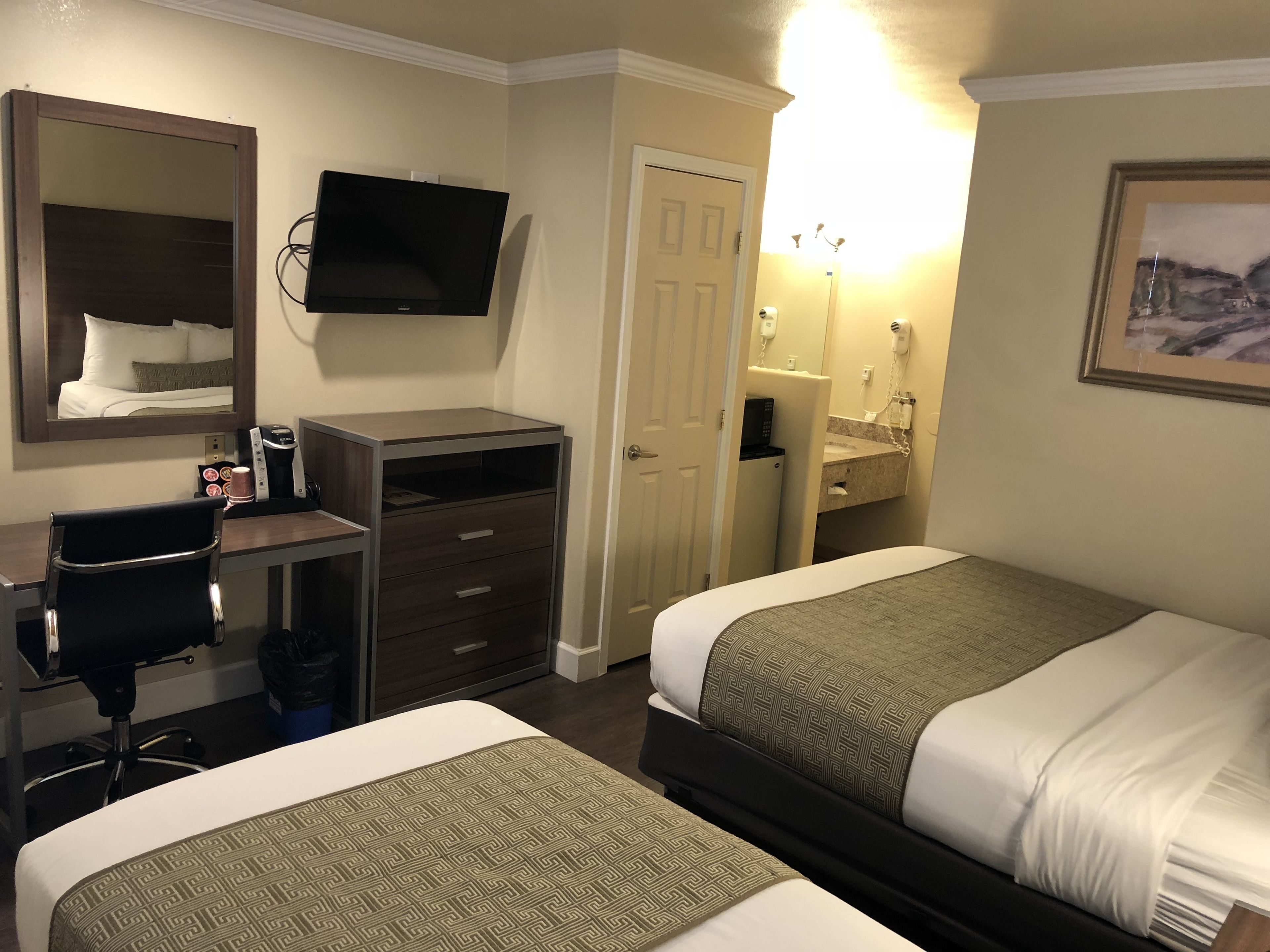 A hotel room with two beds, a desk with a chair, a TV, a mirror, a door, and a bathroom with a sink and mirror.