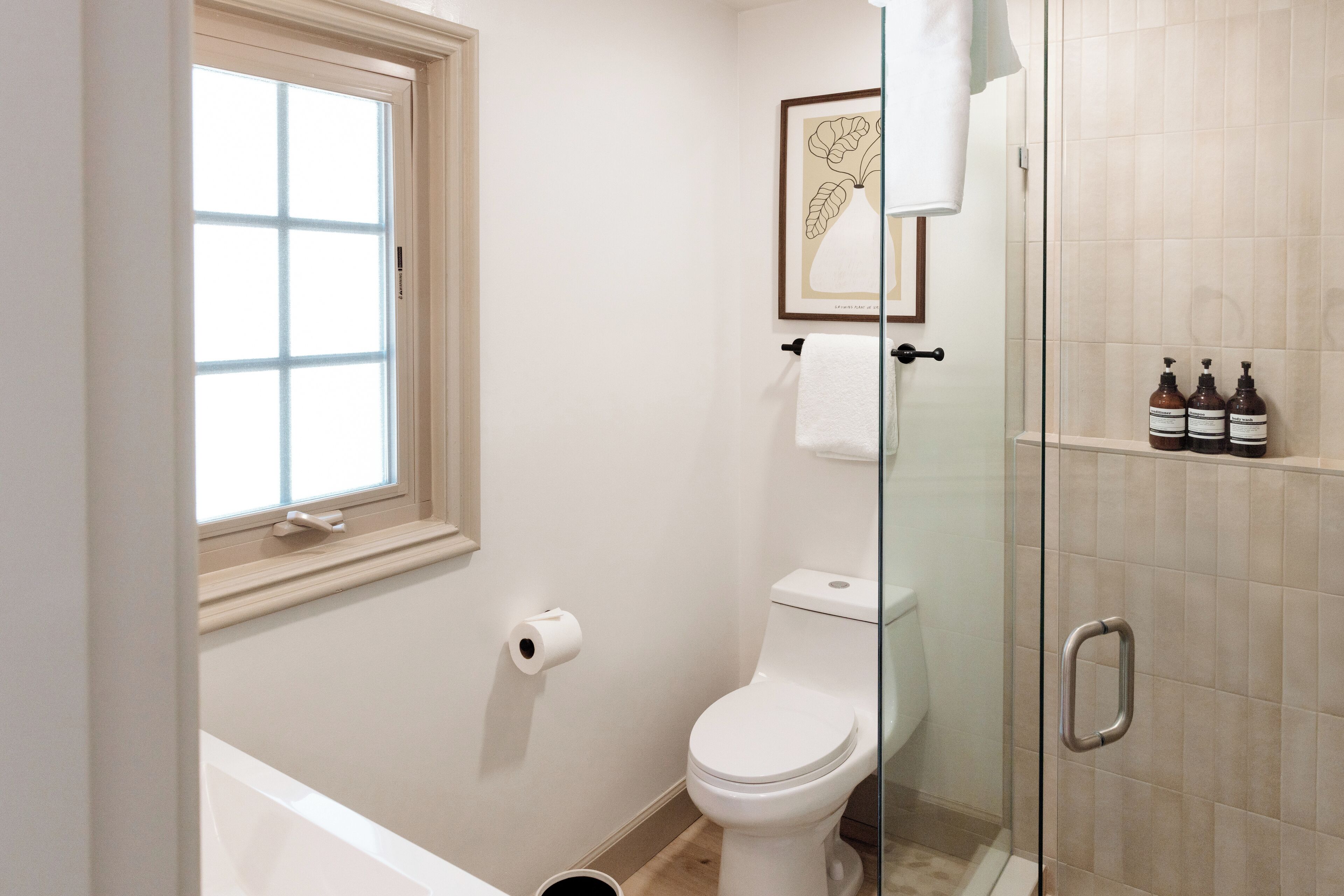 A bathroom with a toilet, a glass shower enclosure, a window, and a framed artwork on the wall.