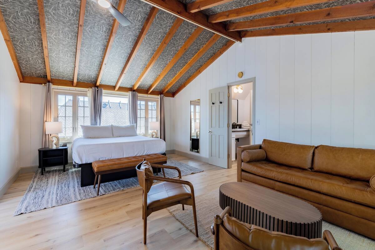 Un dormitorio con cama, piso de madera, un banco, un sofá de cuero marrón y una mesita.