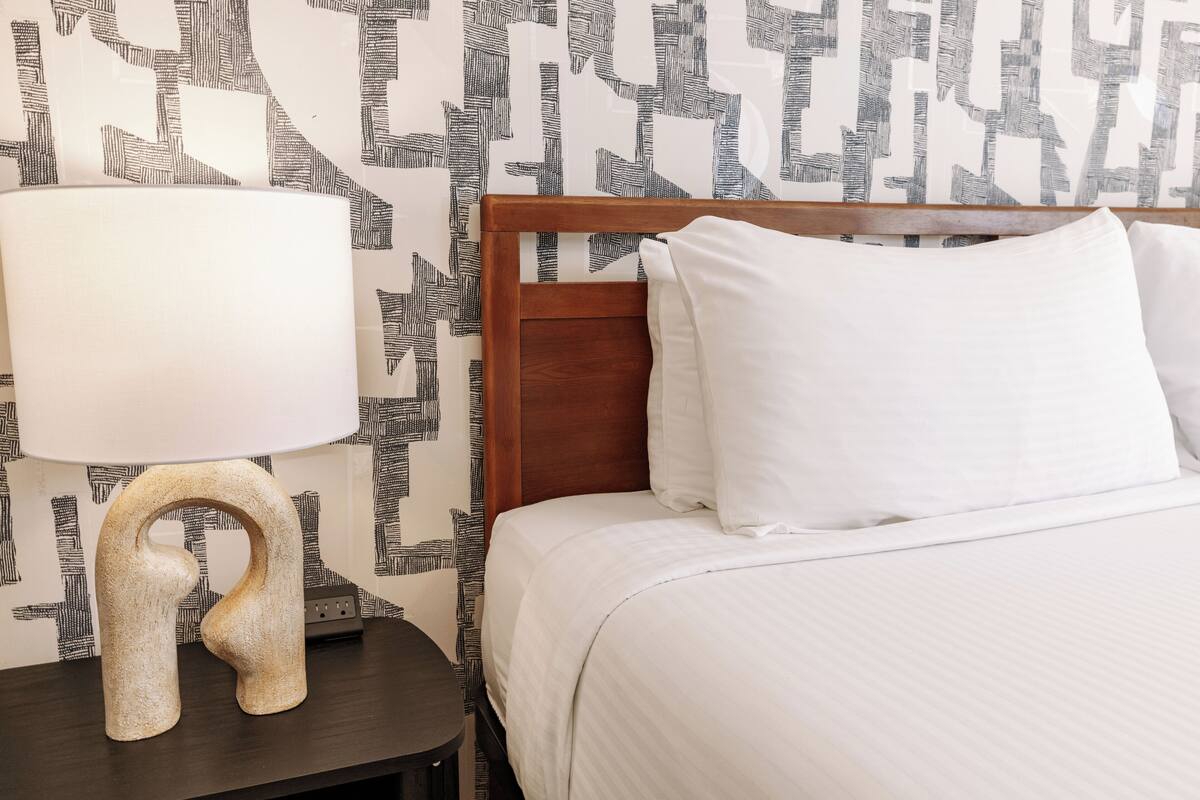 Una cama bien hecha con ropa blanca, un cabecero de madera y una mesita de noche con una lámpara.