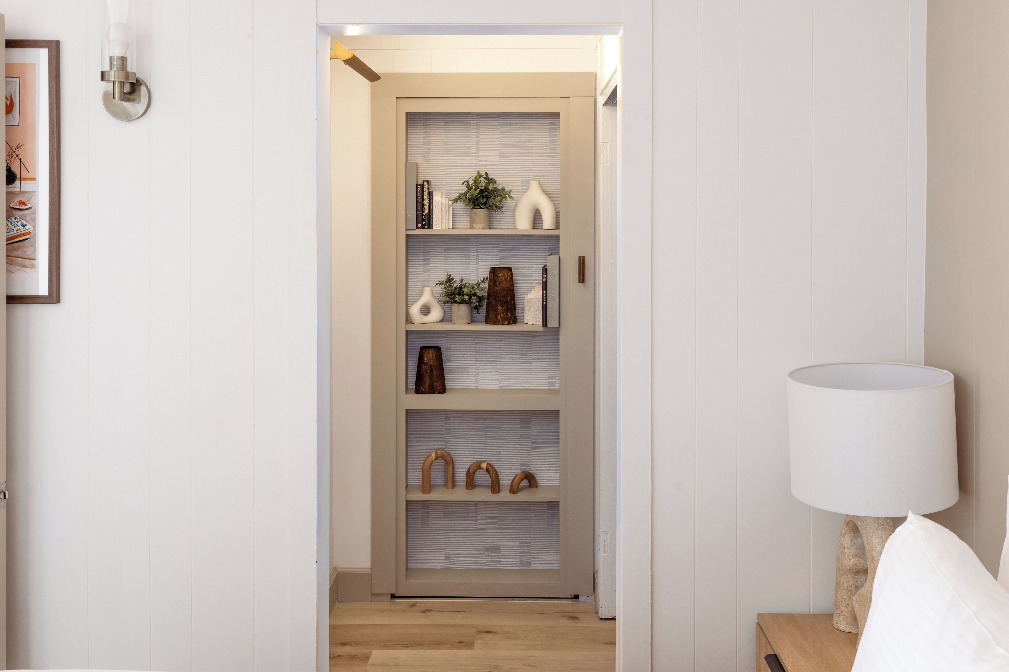 A hallway with a wooden floor, a white wall, and a shelf with decorative items.