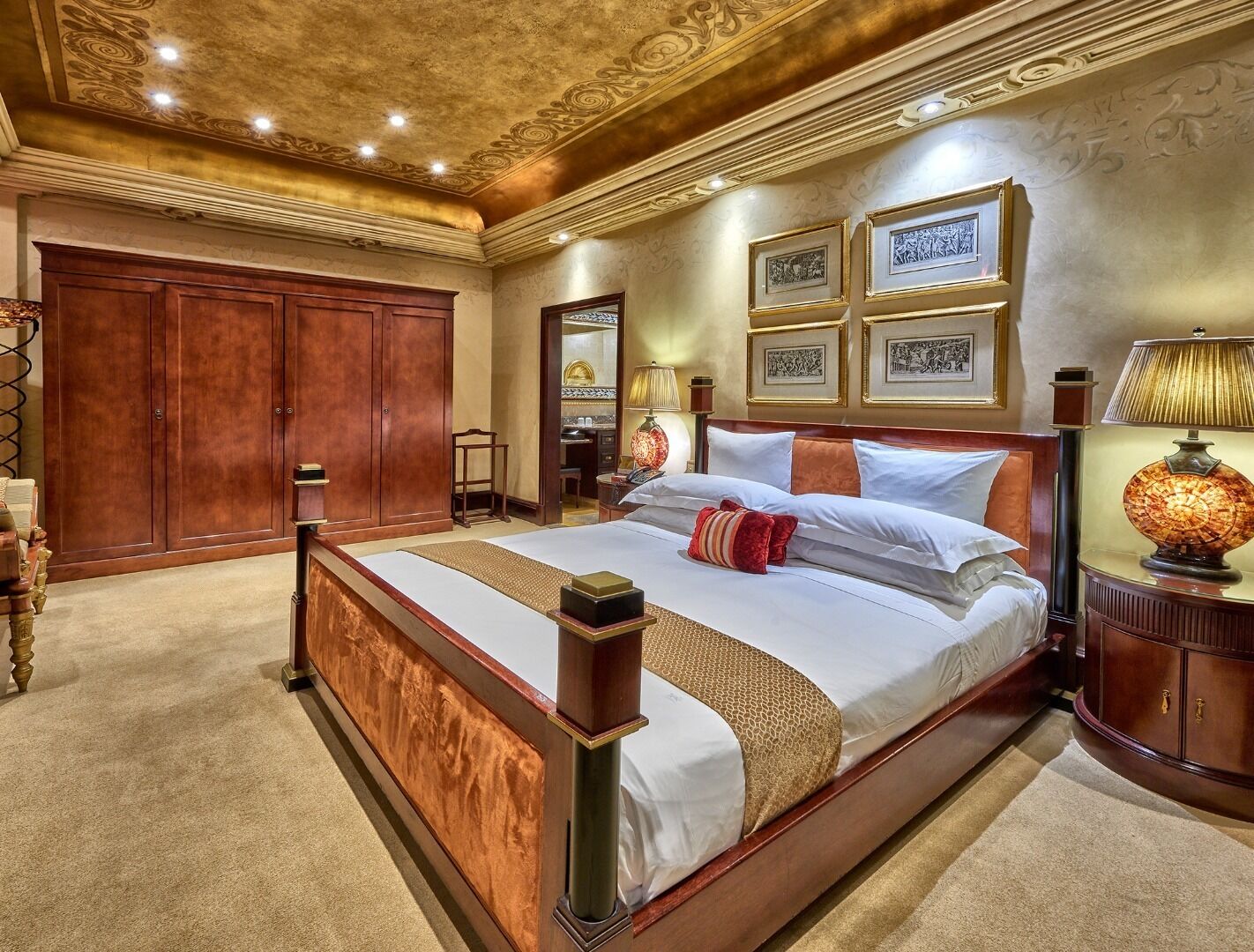 Una cama grande y ornamentada con estructura de madera y muchos almohadones, una mesita de noche con una lámpara, y cuadros en la pared.