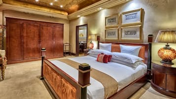 Una cama grande y ornamentada con estructura de madera y muchos almohadones, una mesita de noche con una lámpara, y cuadros en la pared.
