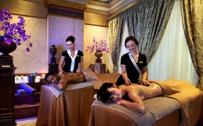 Salão de massagens com duas macas, um vaso de flores roxas e um espaço de bar com copos.