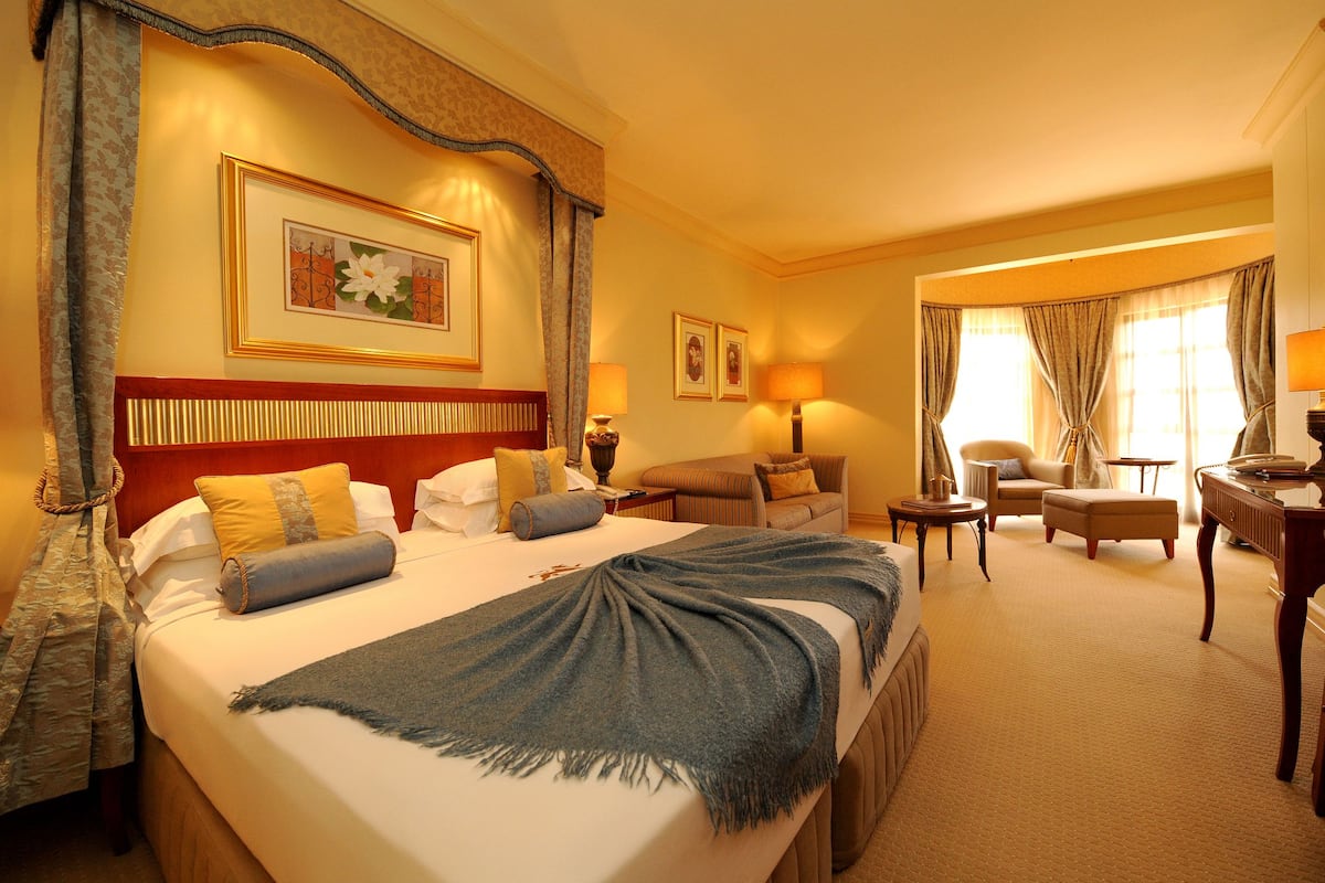 A hotel room with a large bed, a sitting area with a sofa and armchairs, a desk with a lamp, and a window with curtains.