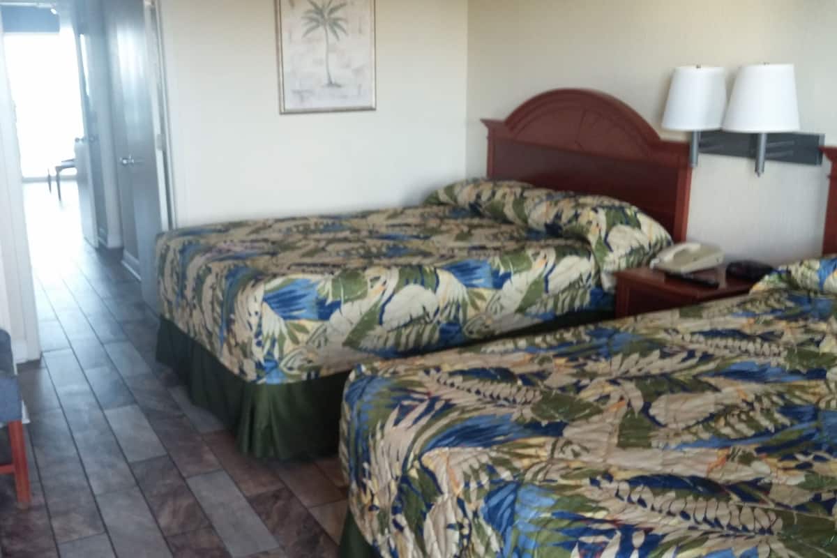 A hotel room with two beds, a chair, a desk with a lamp, and a door leading to a hallway.