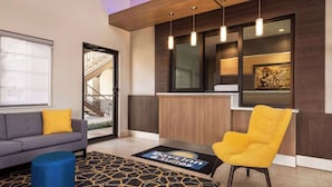 A hotel lobby with a sofa, a yellow armchair, a blue ottoman, a reception desk, and a staircase.