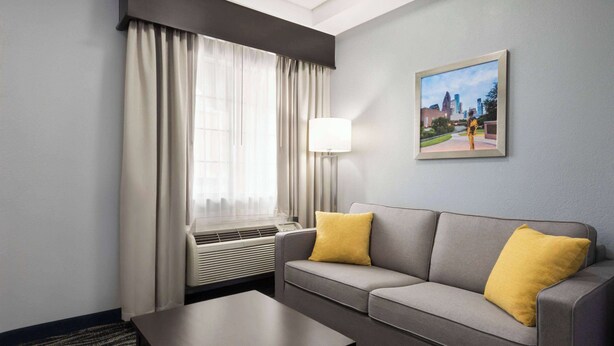 A hotel room with a grey sofa, a yellow pillow, a black coffee table, a window with curtains, an air conditioning unit, and a framed picture on the wall.
