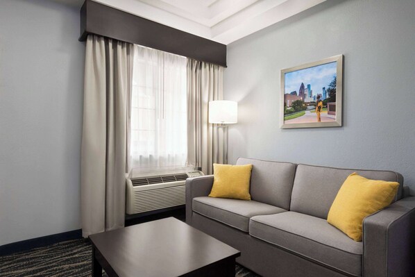 A hotel room with a grey sofa, a yellow pillow, a black coffee table, a window with curtains, an air conditioning unit, and a framed picture on the wall. - Days Inn & Suites by Wyndham Houston Hobby Airport (Houston)