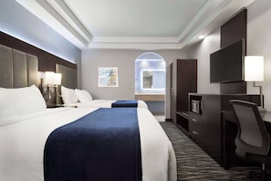 Habitación de hotel con una cama grande, un escritorio, una silla, un televisor y un baño visible a través de un arco.