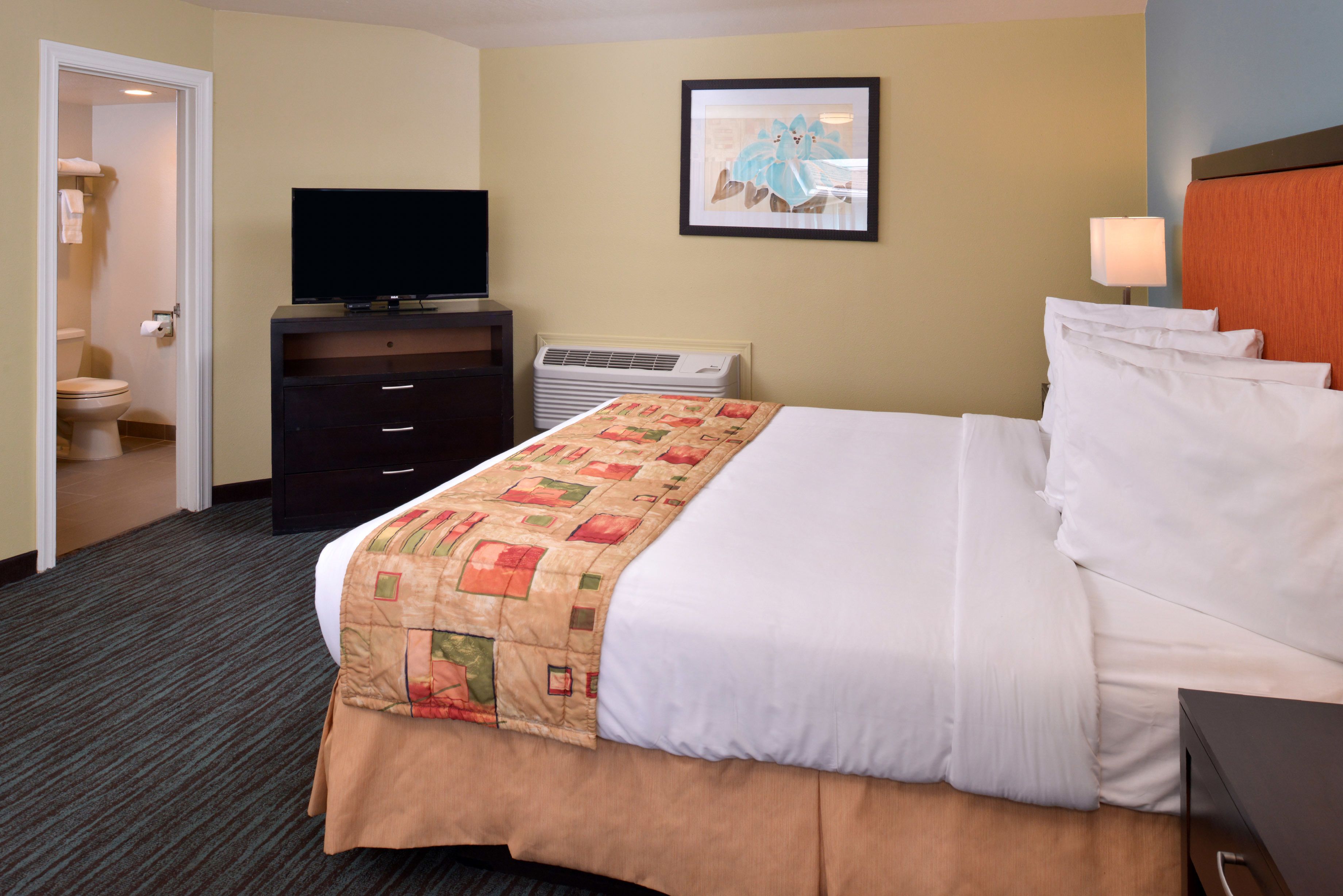 A hotel room with a bed, a television on a dresser, an air conditioning unit, and a painting on the wall.