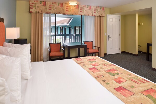 A hotel room with a bed, a desk, two chairs, a lamp, and a view of a building through a window.