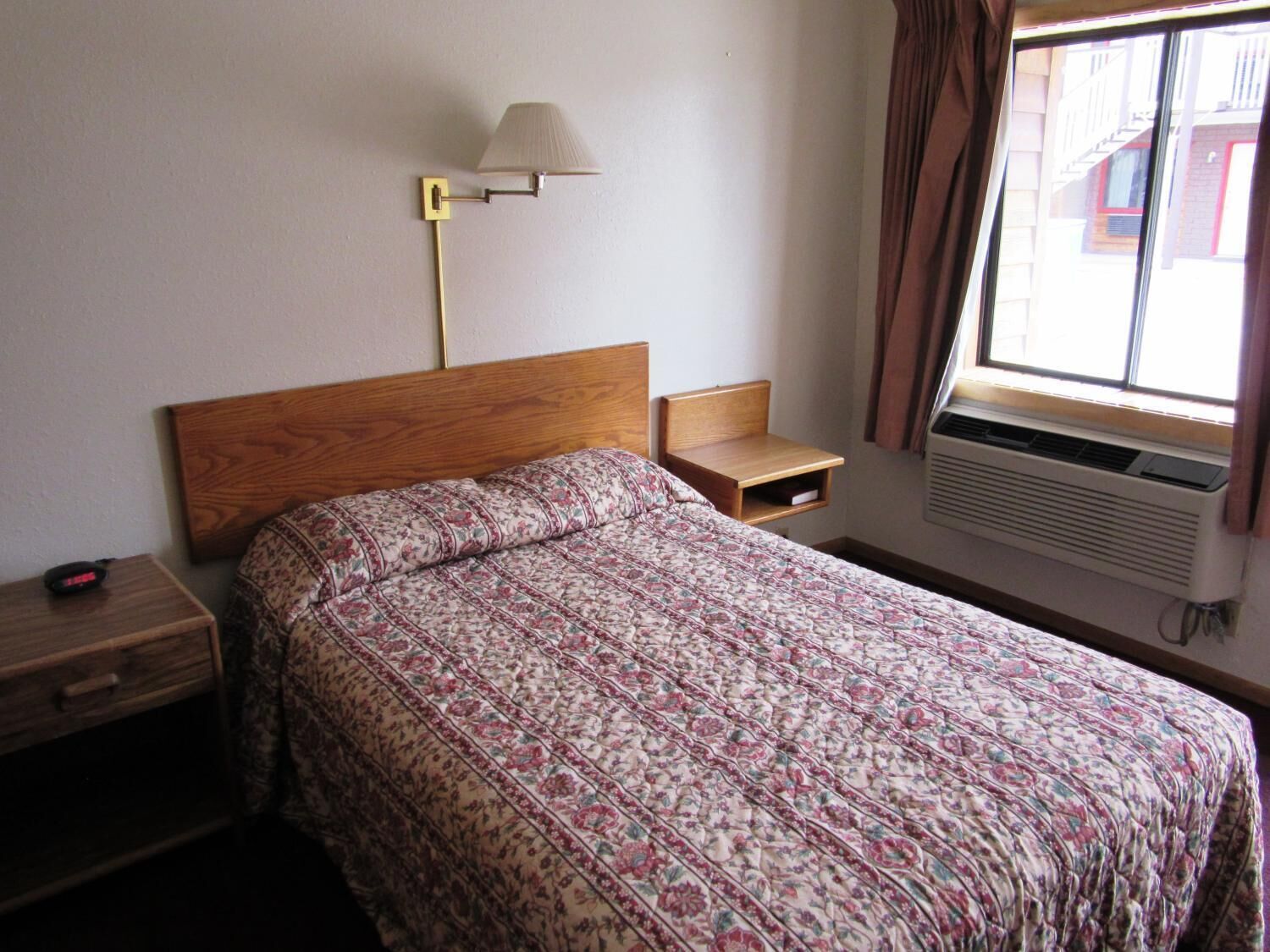 Una camera da letto con una testiera in legno, una coperta a motivi, un comodino con una lampada e una finestra con tende.