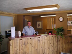 A wooden reception desk with a sign, a framed deer head, and a clock on the wall. - Traveler's Lodge (West Yellowstone)